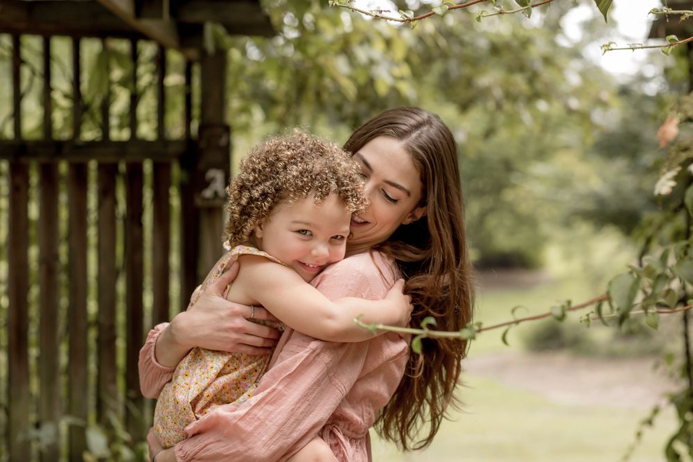 A mother hugs her curly-haired toddler daughter tightly in a peaceful garden setting.