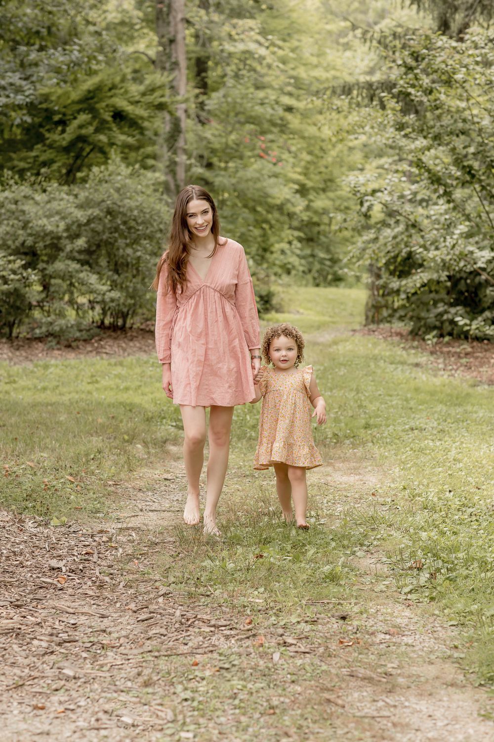 A young mother in pink dress walks hand-in-hand with her toddler daughter on a forest path.