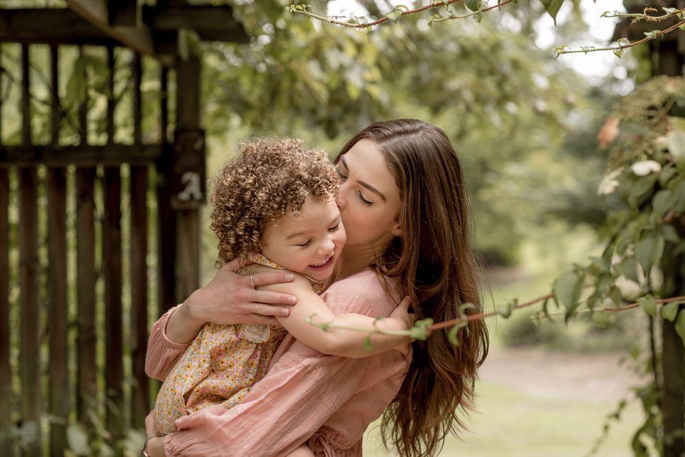 A loving mother kisses her young curly-haired daughter in a lush green outdoor setting.
