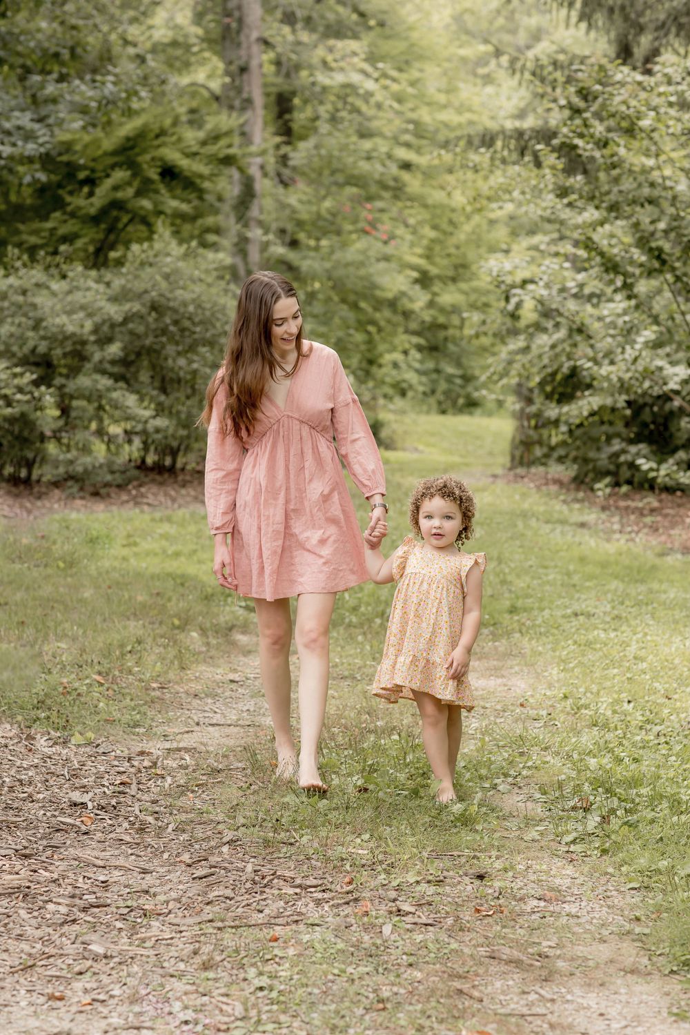 A barefoot mother in a pink dress holds hands with her toddler daughter on a garden path.