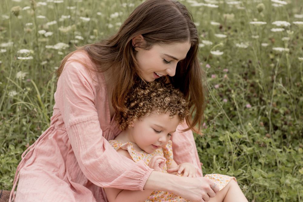 Mother in pink dress tenderly embracing her curly-haired toddler in a wildflower meadow.