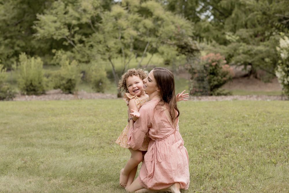A mother in a pink dress kisses her curly-haired toddler girl outdoors in a green park.