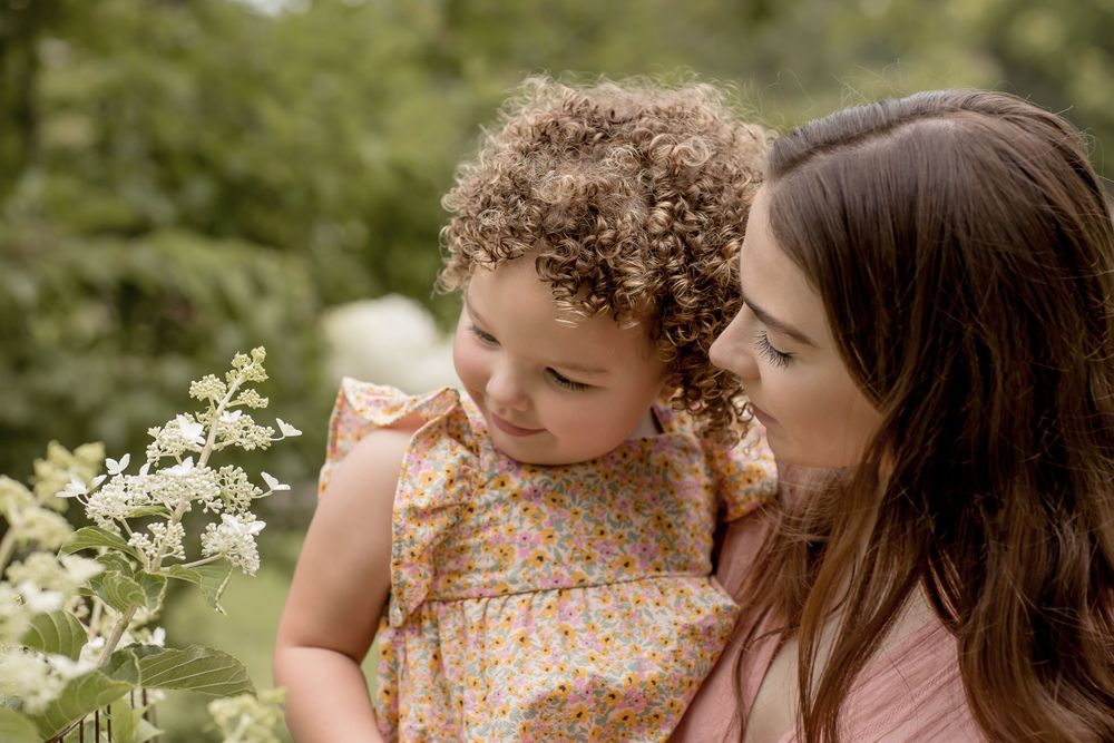 A mother tenderly holds her curly-haired toddler near white wildflowers in a garden.