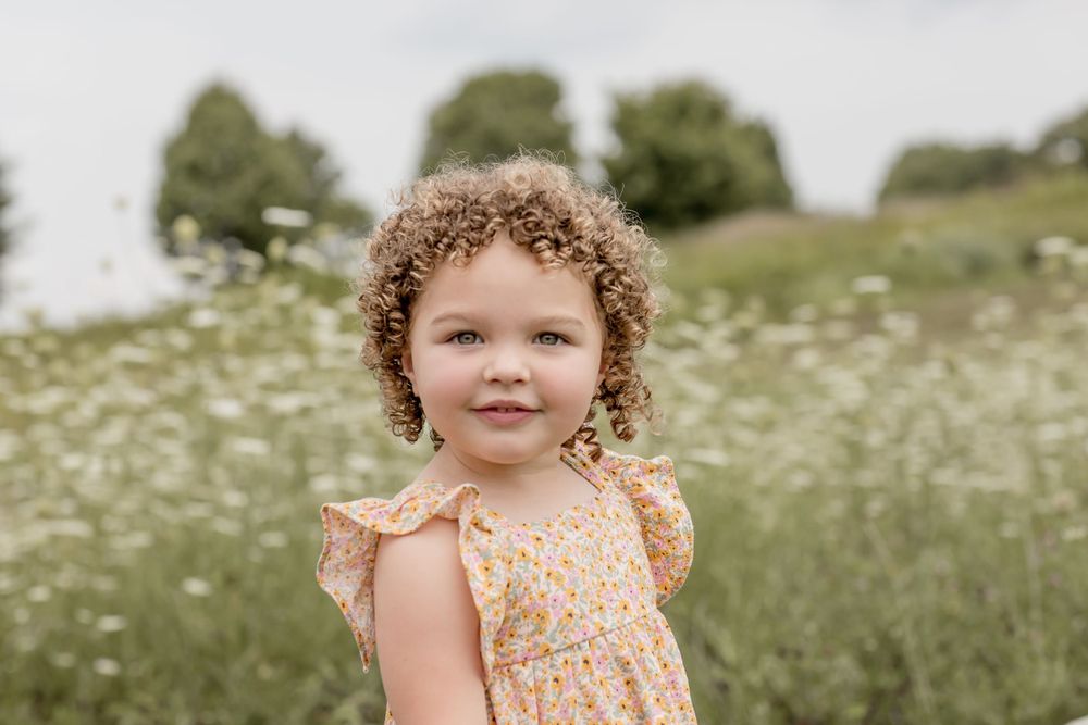 Adorable curly-haired little girl in a floral dress smiling in a wildflower field.
