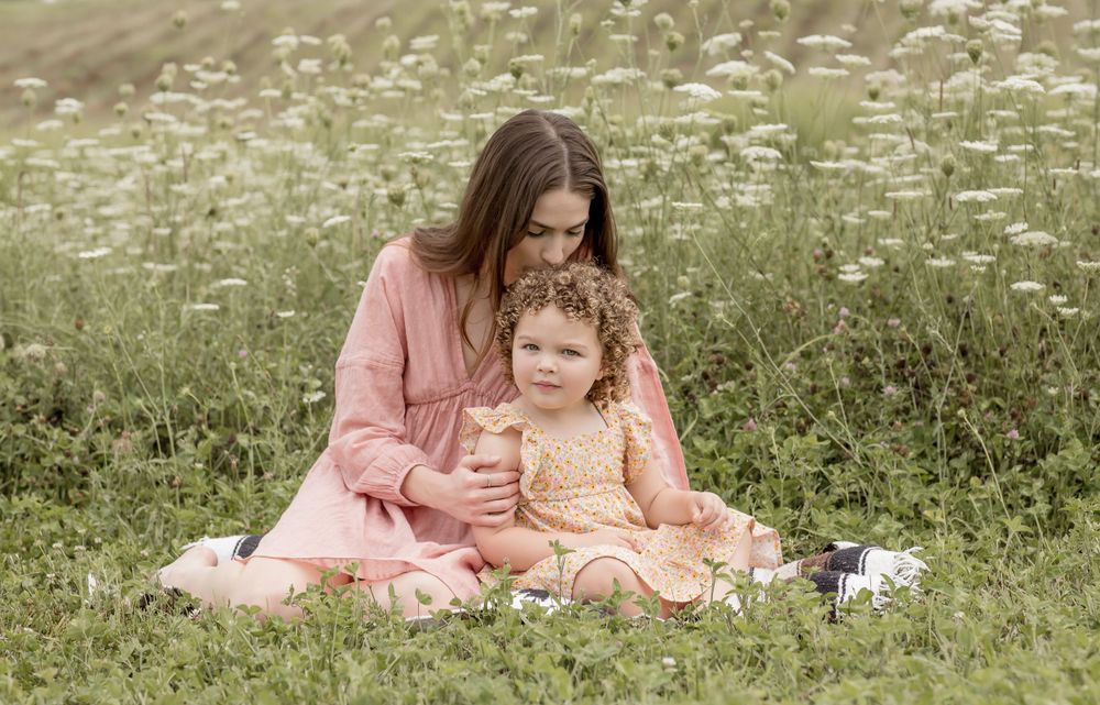 A mother and curly-haired toddler sit together in a blooming white wildflower meadow.