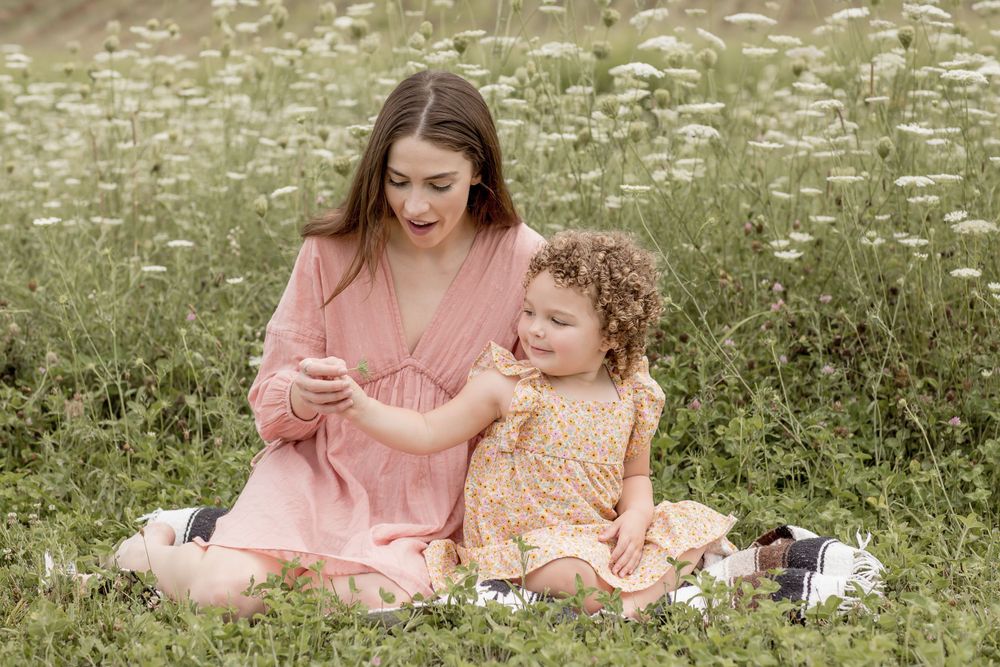A mother in pink dress sits in a wildflower meadow sharing a flower with her toddler.