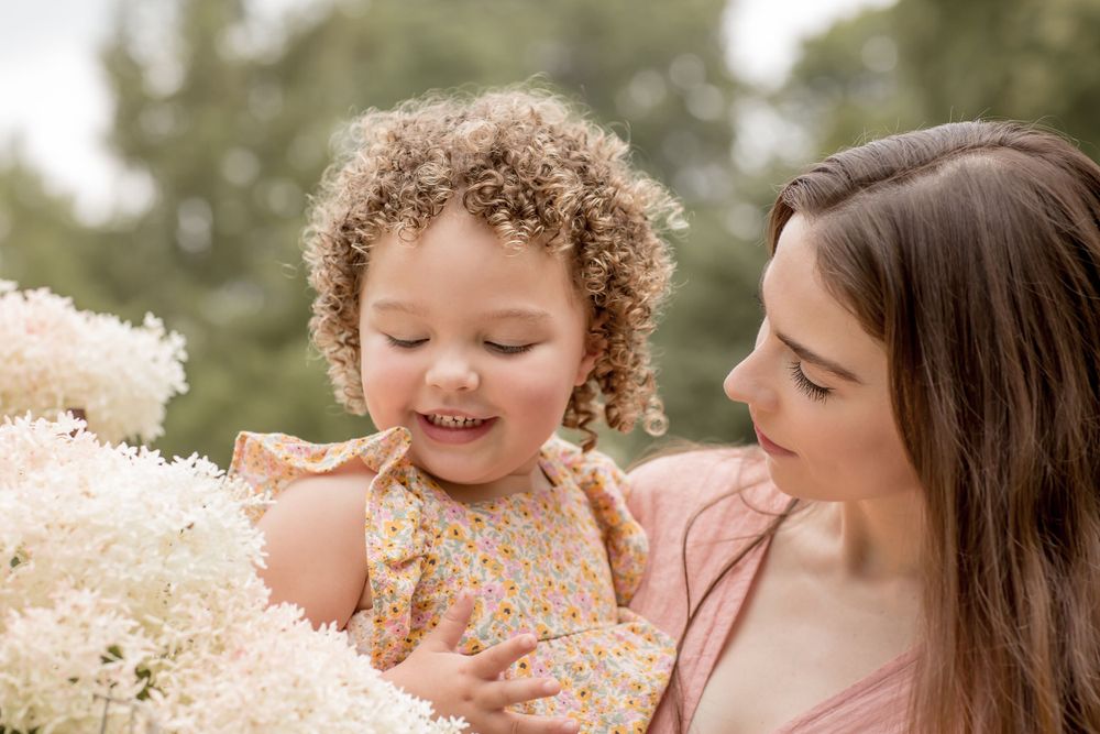 Mother in pink dress holding curly-haired toddler near white hydrangea blooms outdoors.