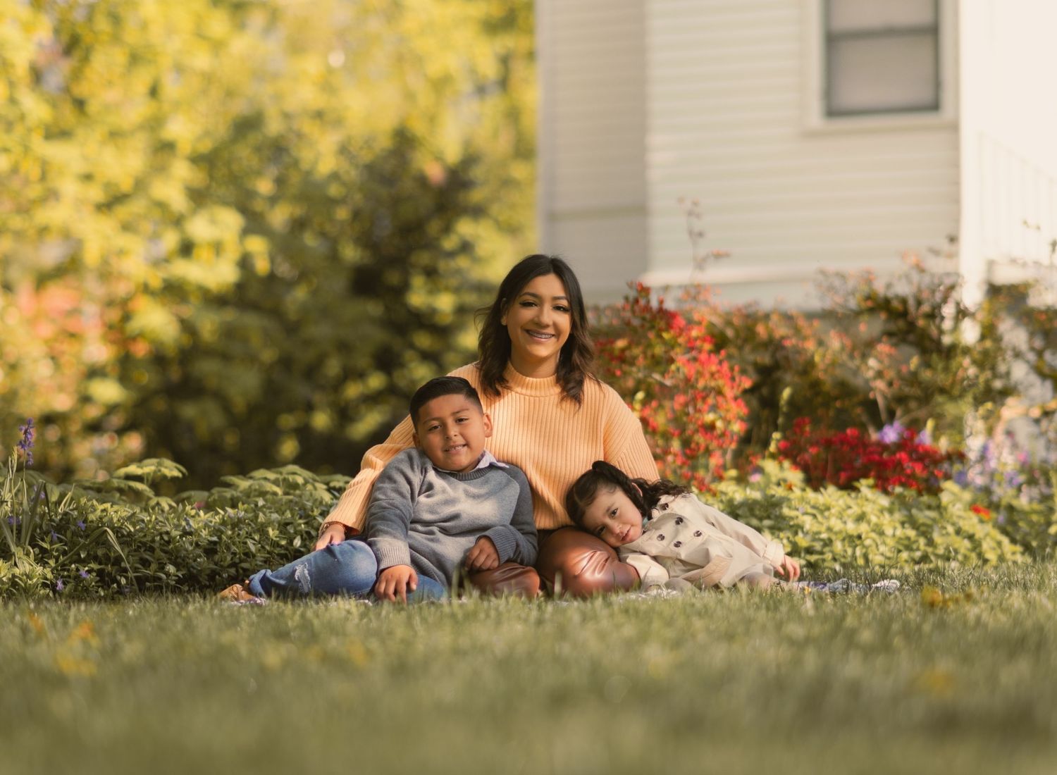 hispanic family of 3 sitting on ground with two kids leaning on mom.