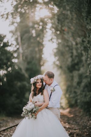 Dreamy forest wedding portrait with couple in white dress and suspenders under sunlit trees.