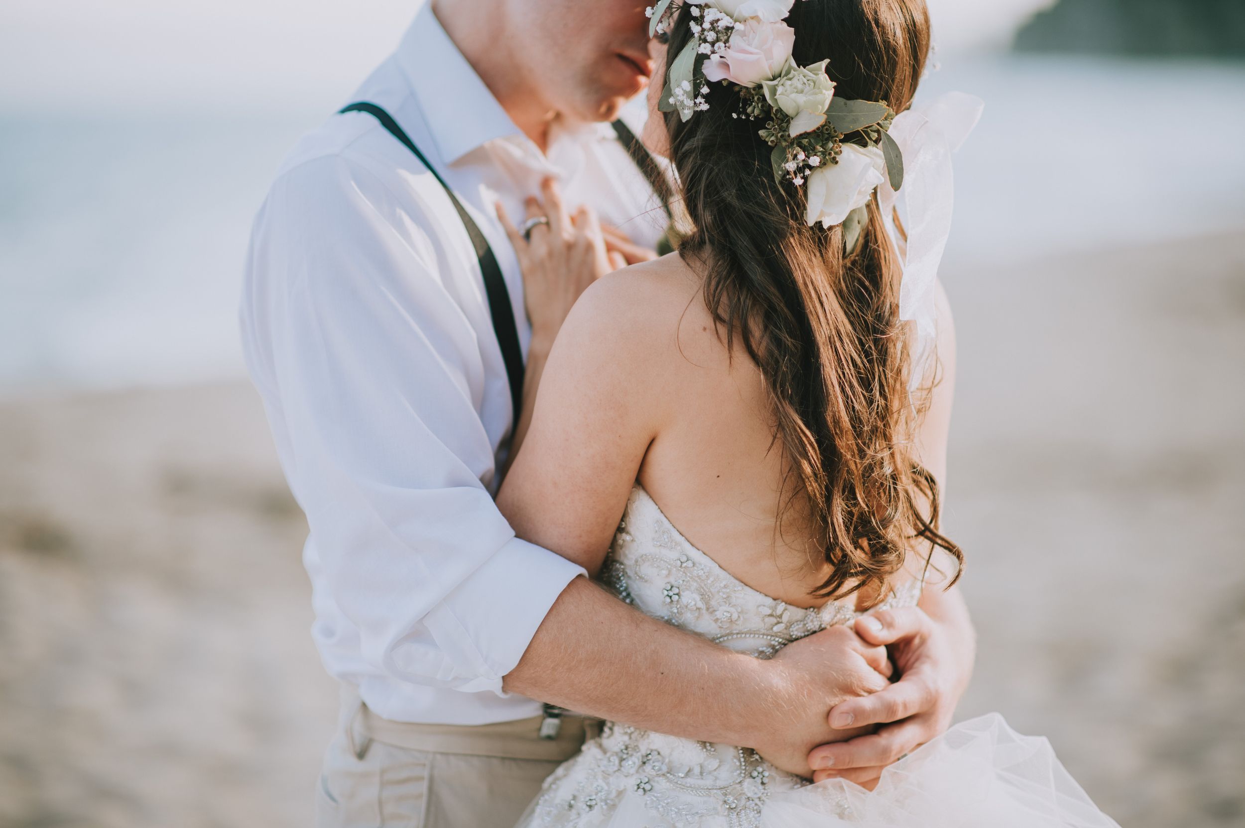 A couple embraces on a beach in casual wedding attire with floral crown details.