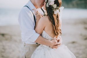 Elegant couple in white embracing on beach with floral details and suspenders.