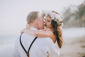 Intimate beachside wedding moment with couple in white and floral crown embracing.