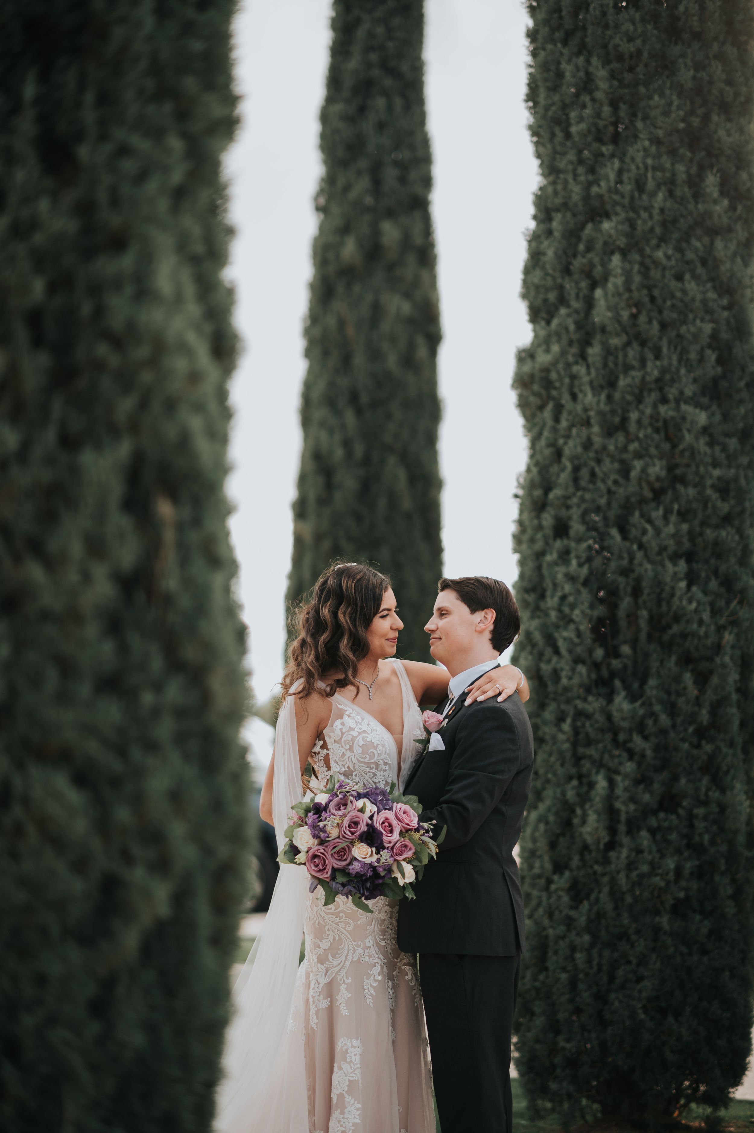 A couple stands between tall cypress trees holding a purple bouquet.