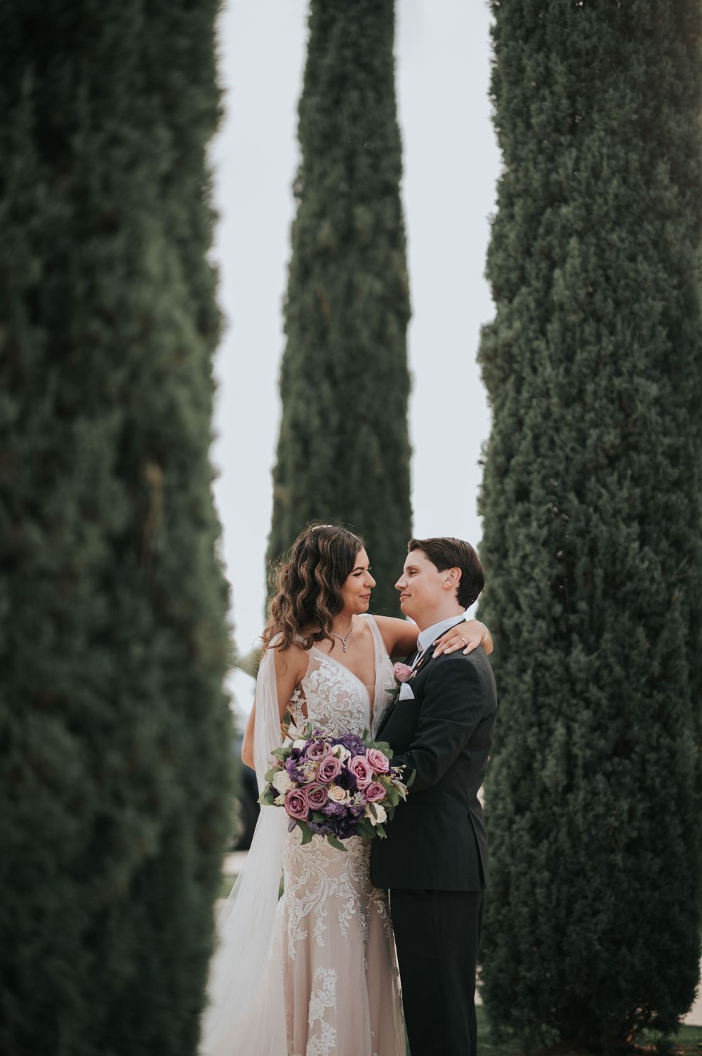 A couple stands between tall cypress trees holding a purple bouquet.