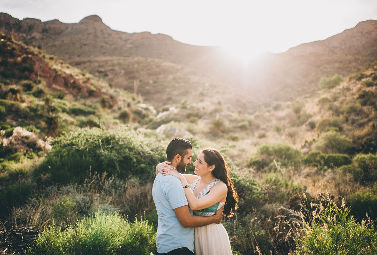 Couple embracing on hillside trail with glowing sunset behind desert mountains.