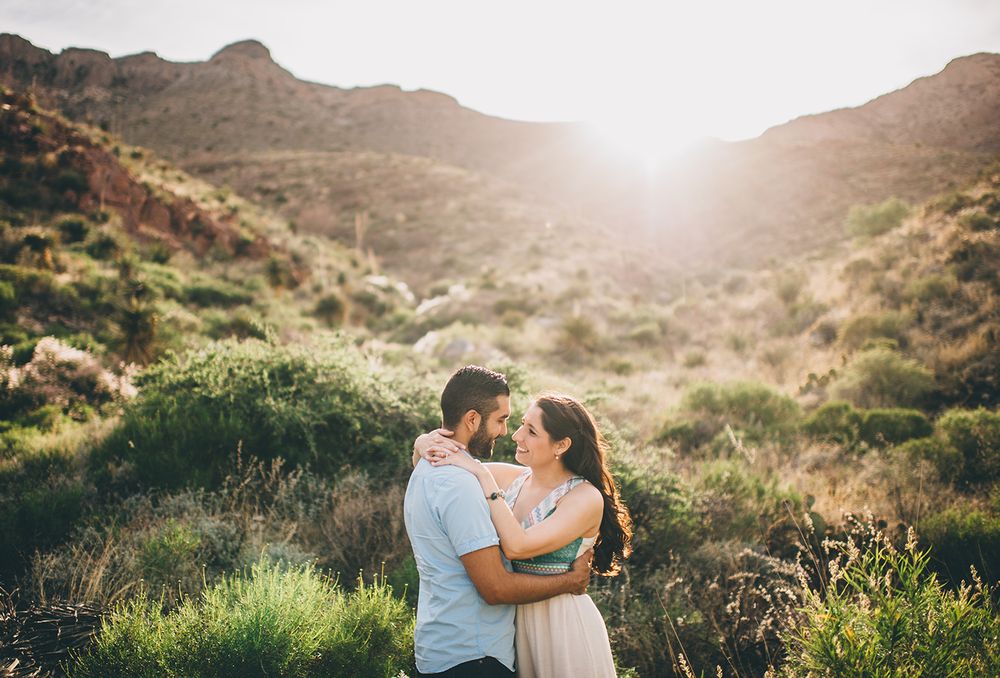 Couple embracing on hillside trail with glowing sunset behind desert mountains.
