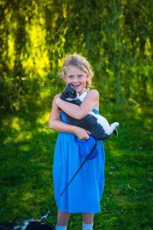 A young child in a blue dress holds a gray and white kitten in a lush garden setting.