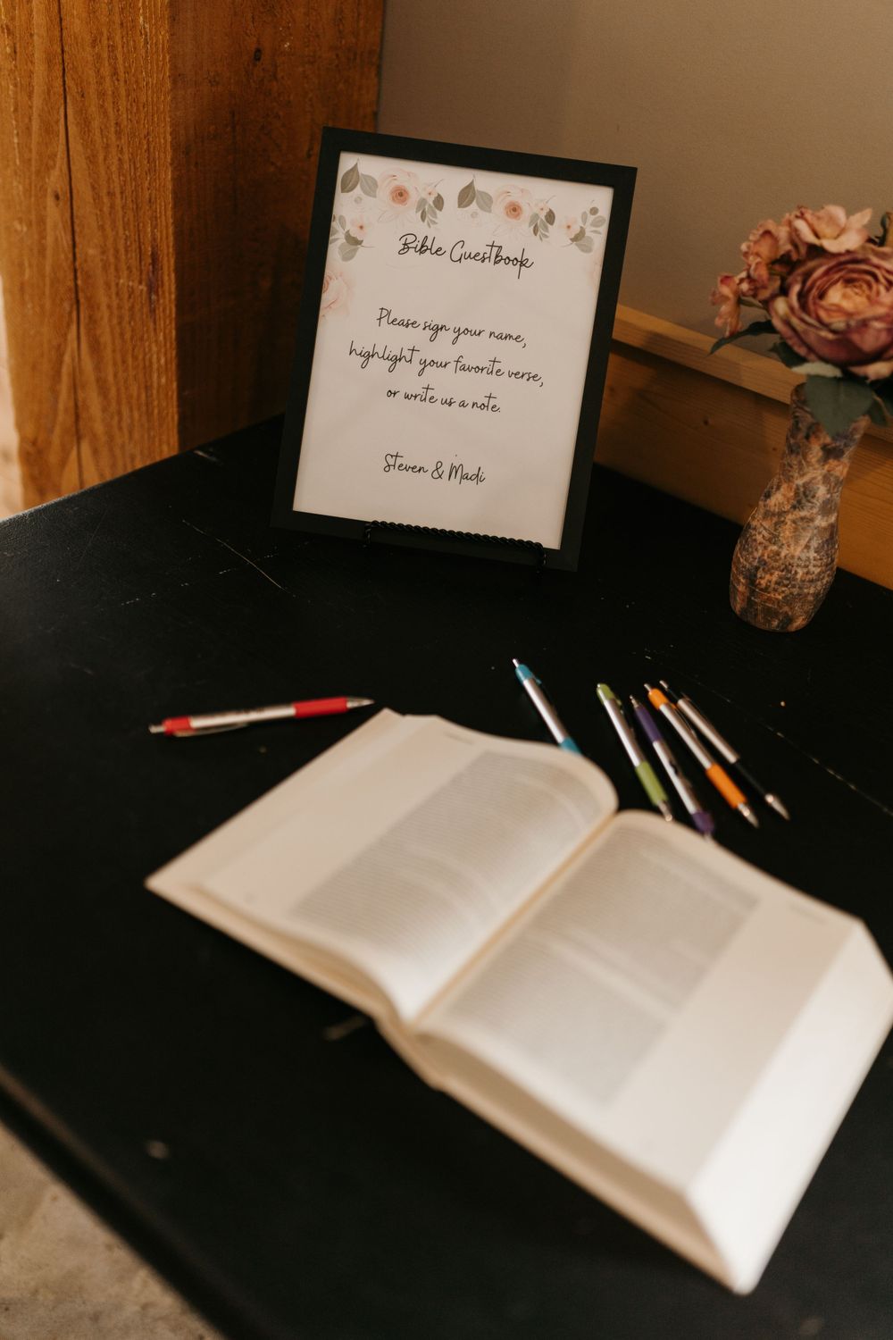 Bible guestbook station with open book, colored pencils, framed sign, and dried floral arrangement.