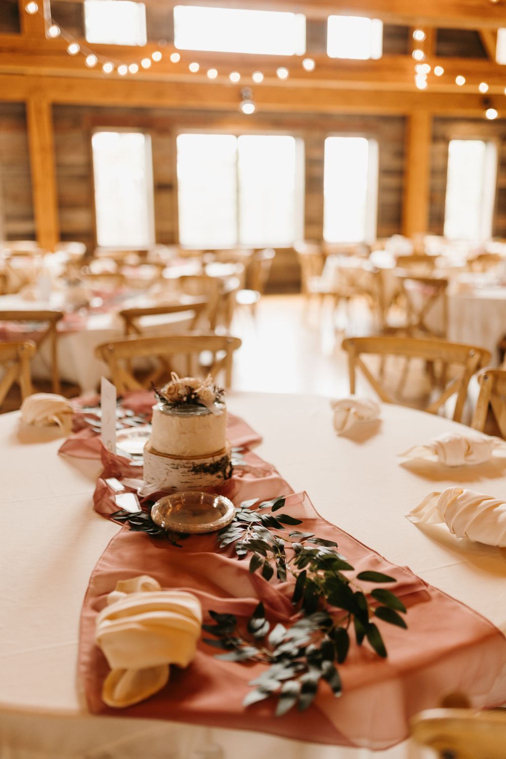 Rustic barn wedding reception head table with dusty rose runner, mini cake, and greenery garland.
