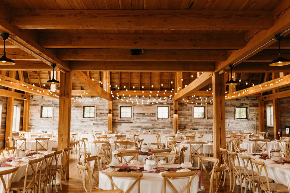 Warm barn wedding venue with exposed wood beams, string lights, and round tables with white linens.