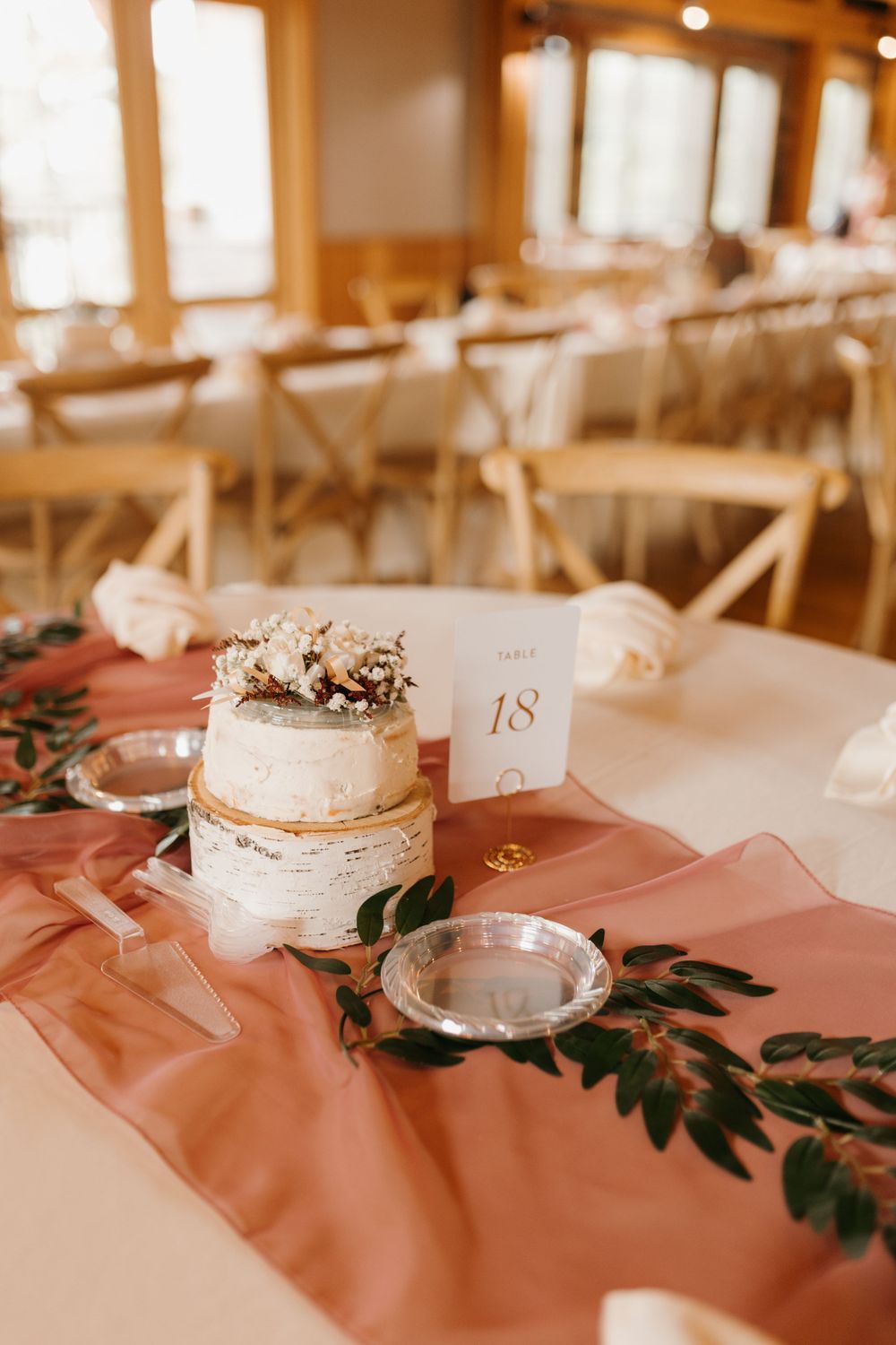 Elegant two-tier naked wedding cake centerpiece on dusty rose runner with greenery in a warmly lit reception hall.