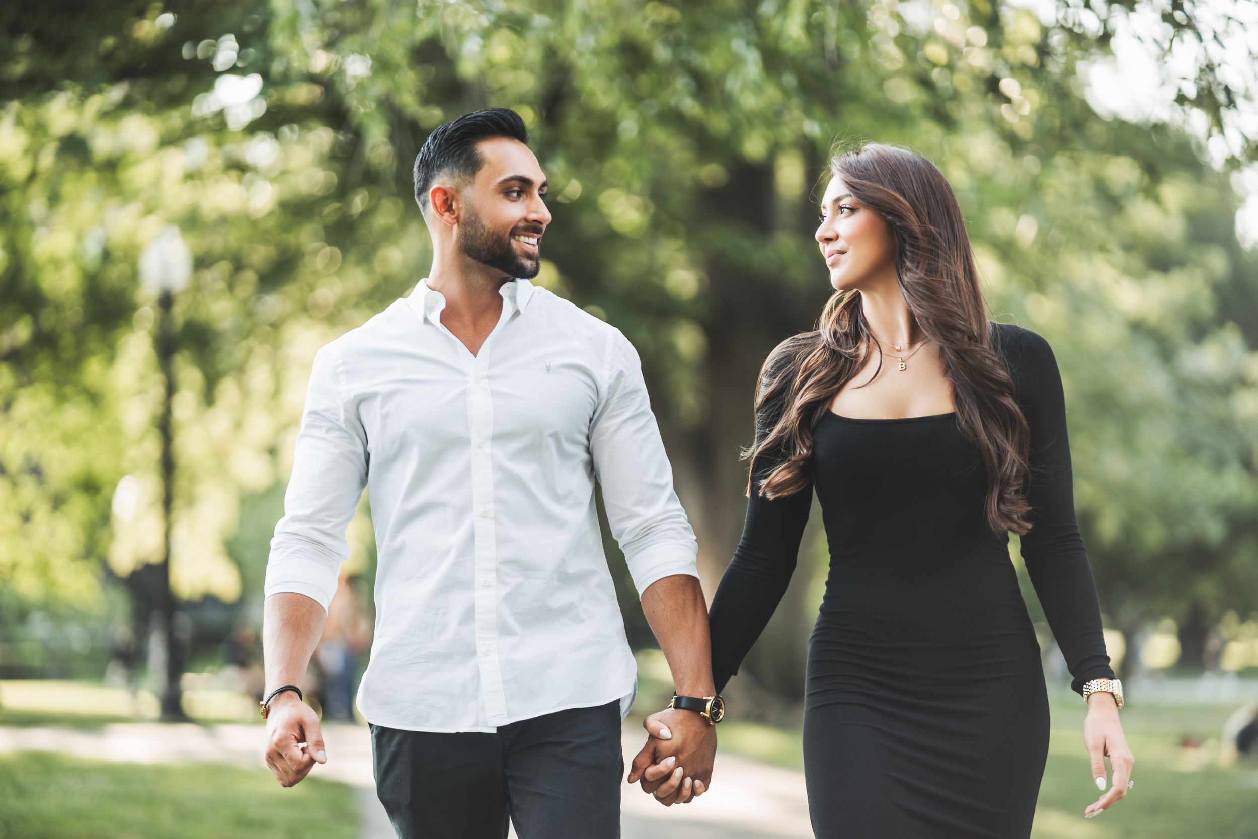 A stylish couple walks hand in hand through a park in elegant attire on a sunny day.