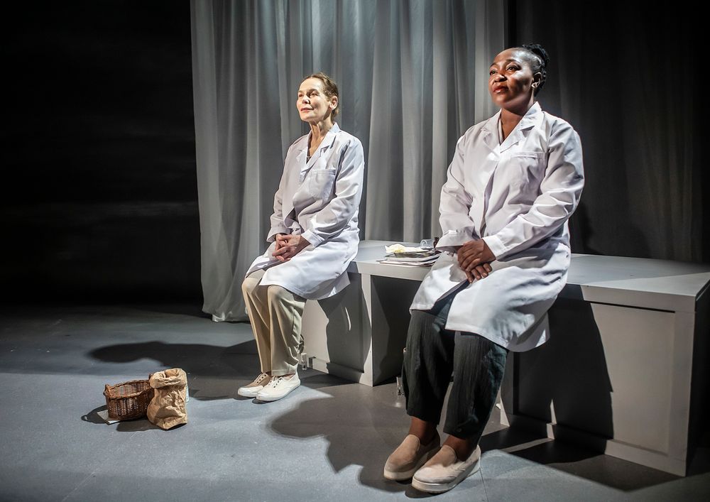 Two performers in white medical coats sit on stage during a theatrical scene.
