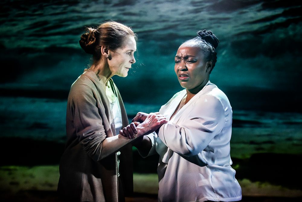 Two performers in dialogue during emotional stage scene with atmospheric blue-green lighting.