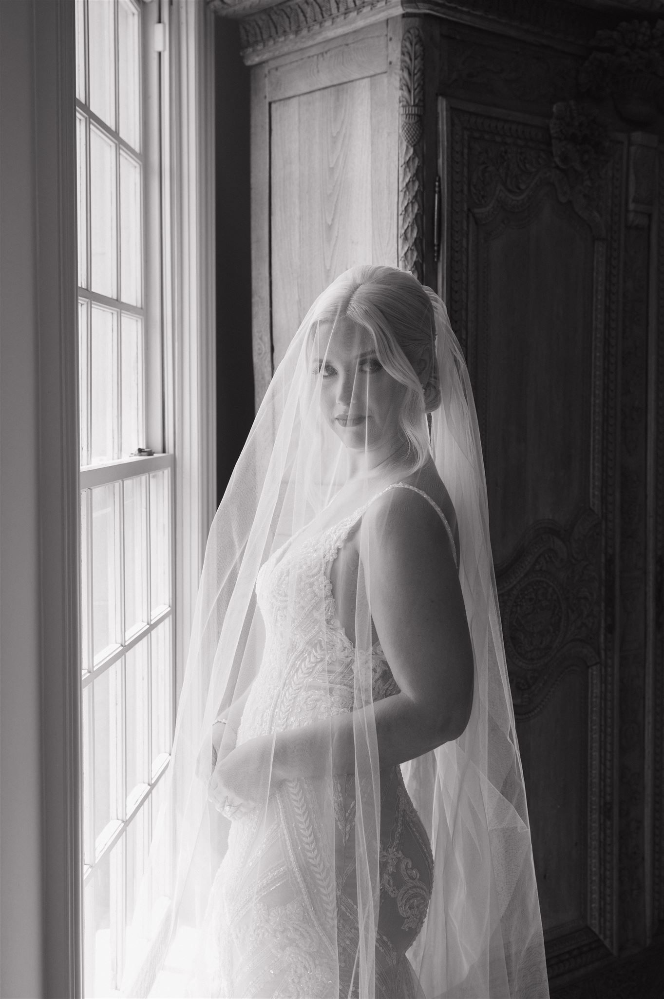 Classic black and white bridal portrait with window light illuminating intricate lace dress and flowing veil.