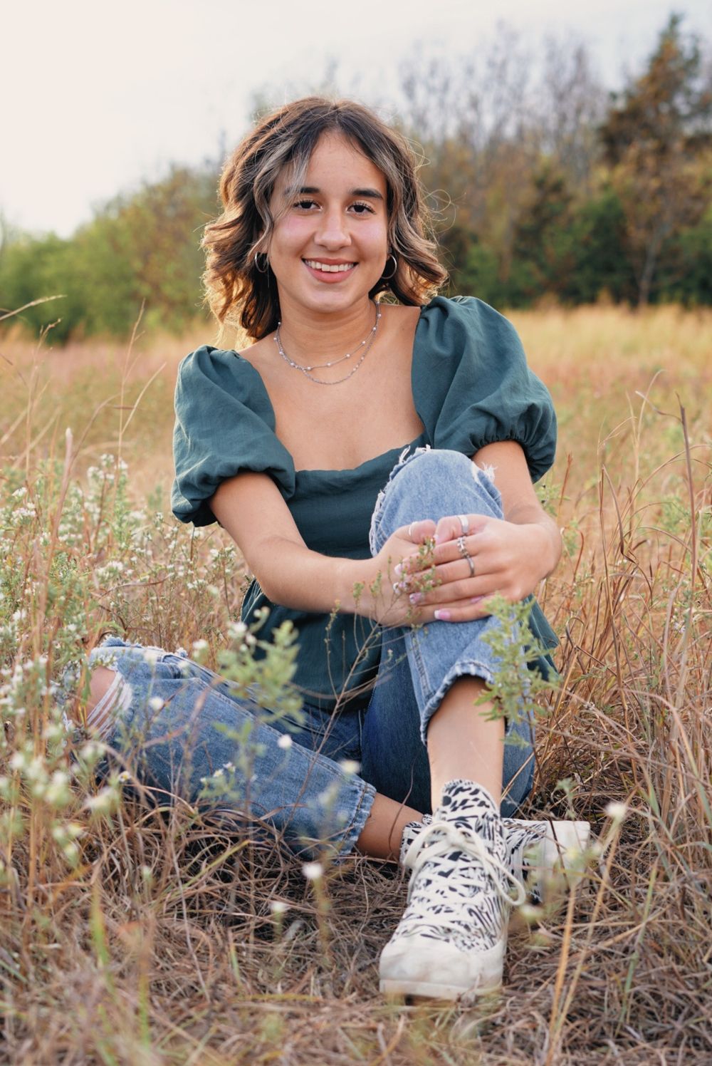 Person in teal top and jeans sitting in tall grass field wearing sneakers.