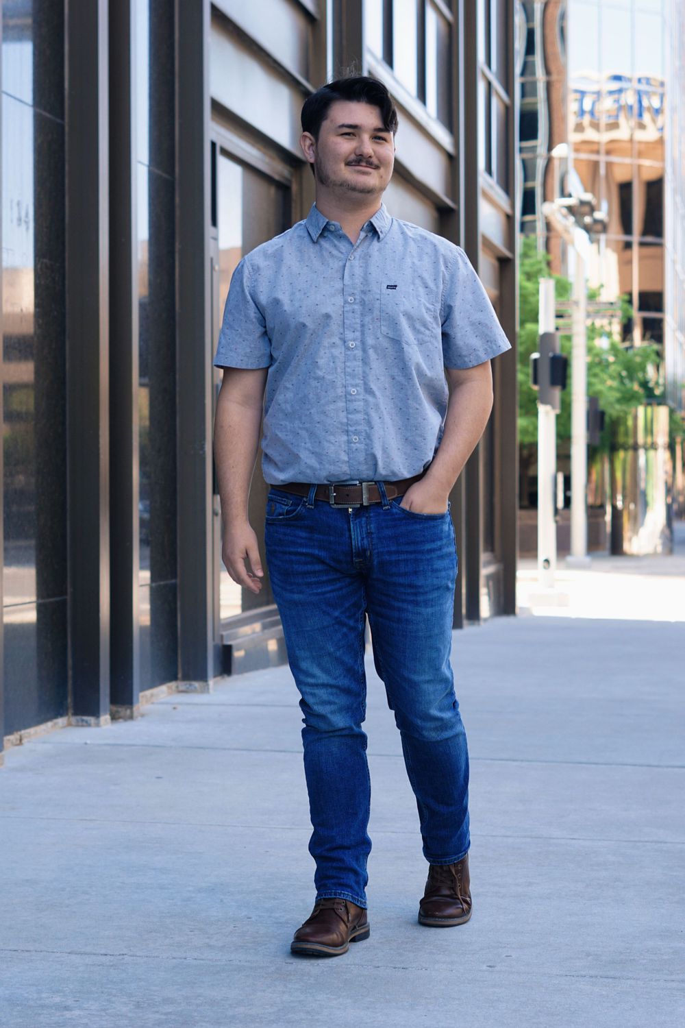 Casual portrait of someone in light blue shirt and jeans standing on city sidewalk.