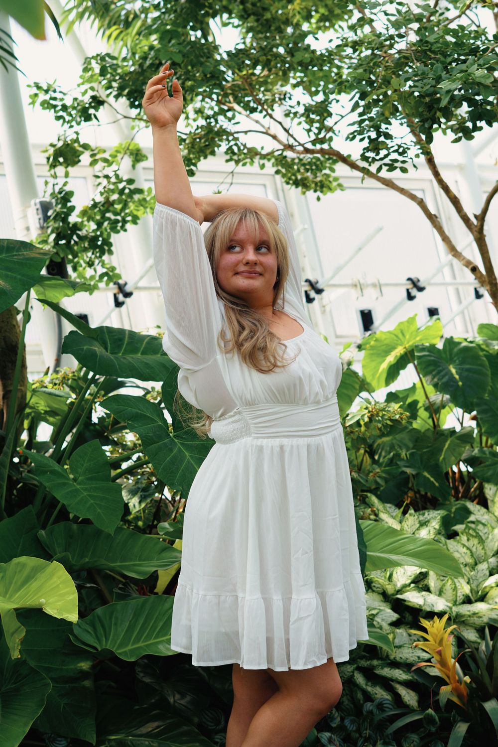 Woman in white sundress poses playfully in a lush greenhouse setting.