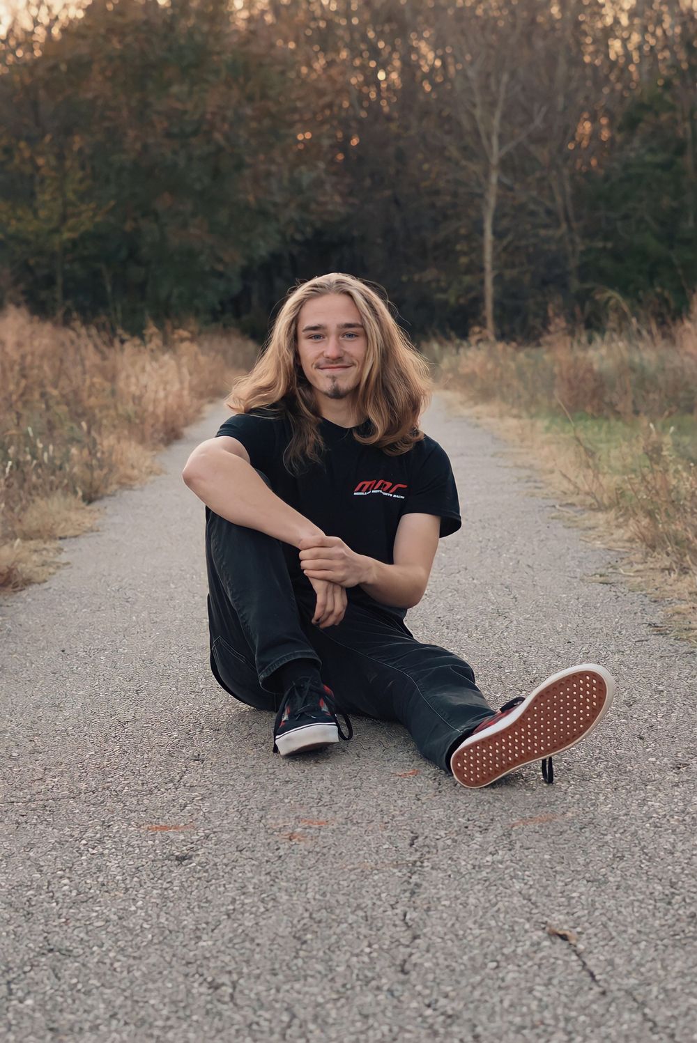 Person sitting on paved path wearing black t-shirt and jeans with casual sneakers at sunset.