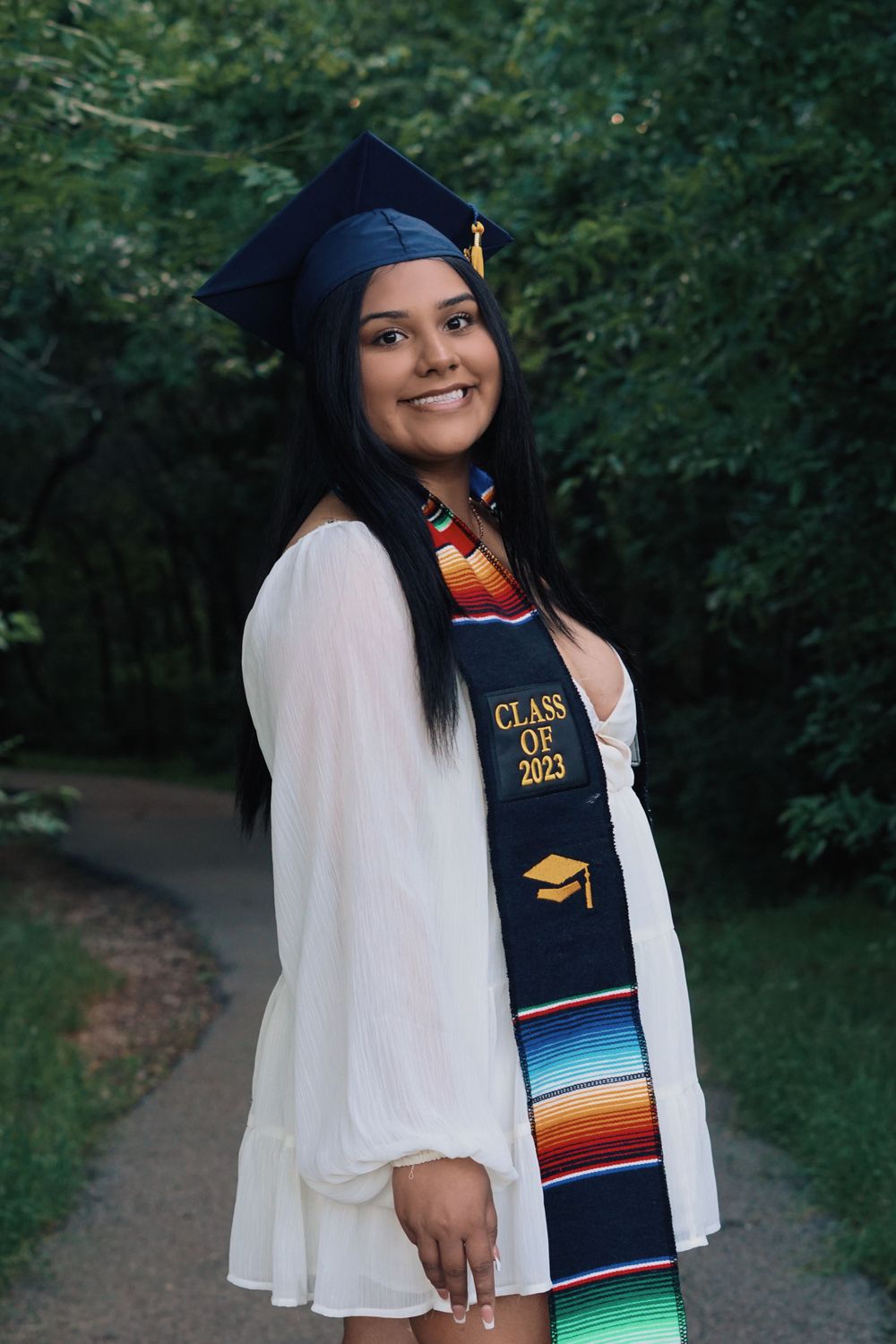 Graduate in white dress and cap with serape stole poses on outdoor path.