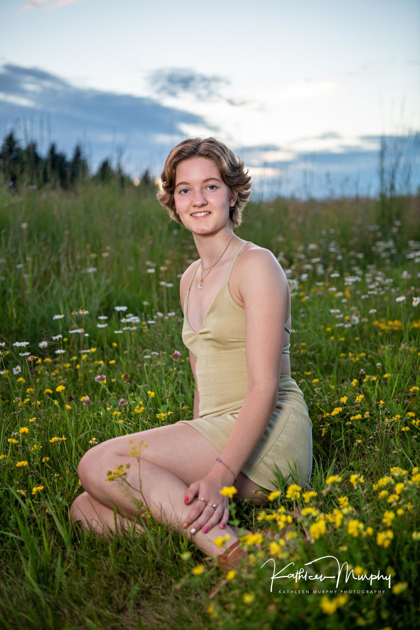 Summer portrait in a beige dress sitting among wildflowers in a mountain meadow at dusk.