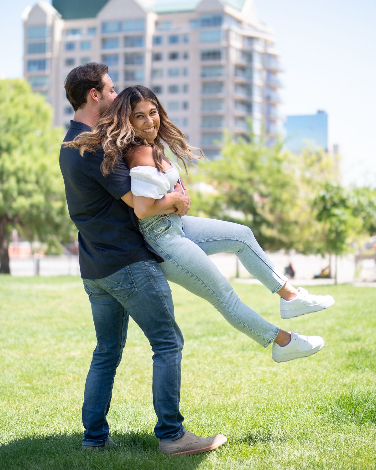 A playful couple shares a tender moment on a grassy lawn with city buildings in the background.