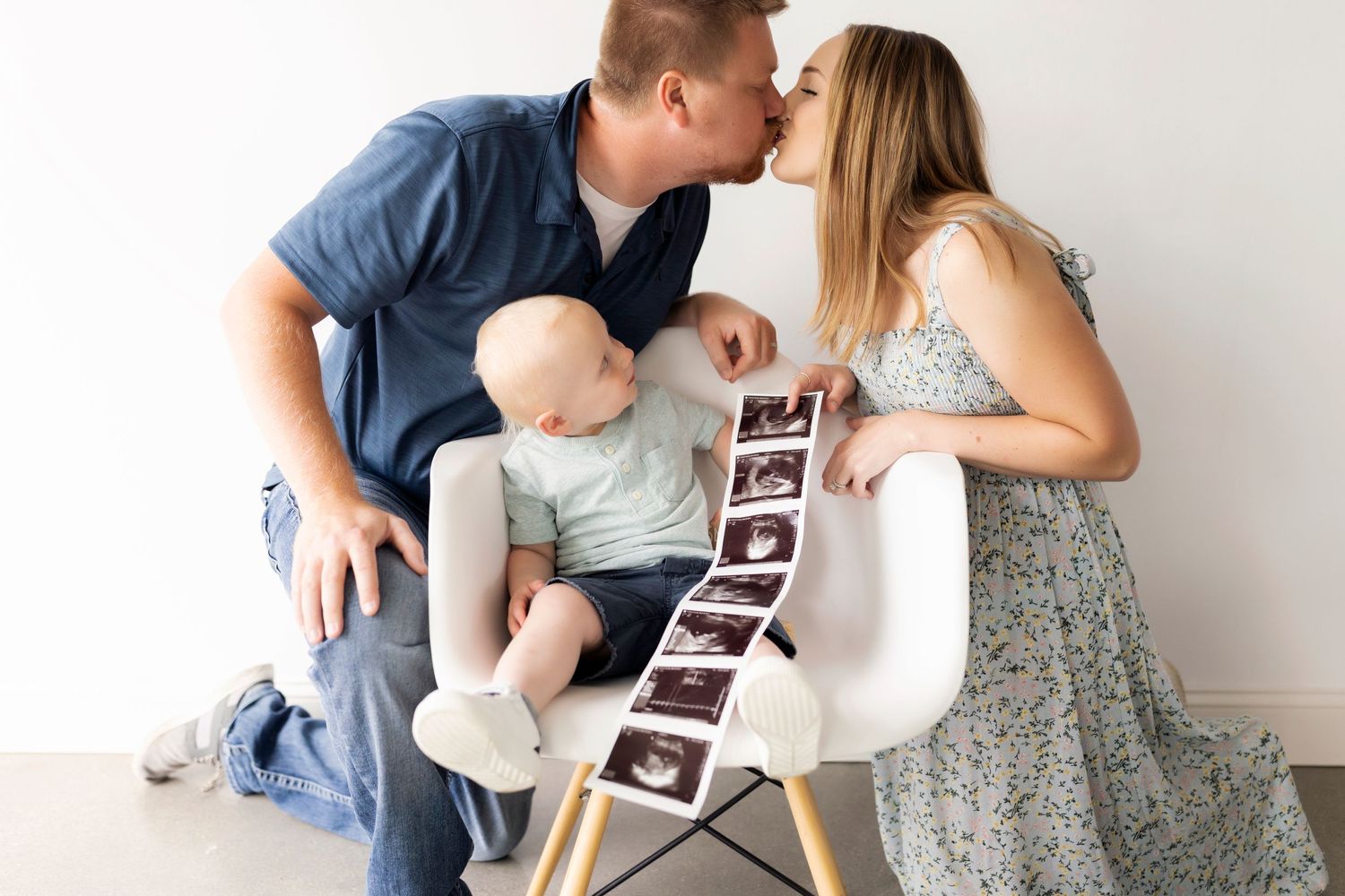 Family sharing intimate moment with baby in white chair while holding ultrasound photos.