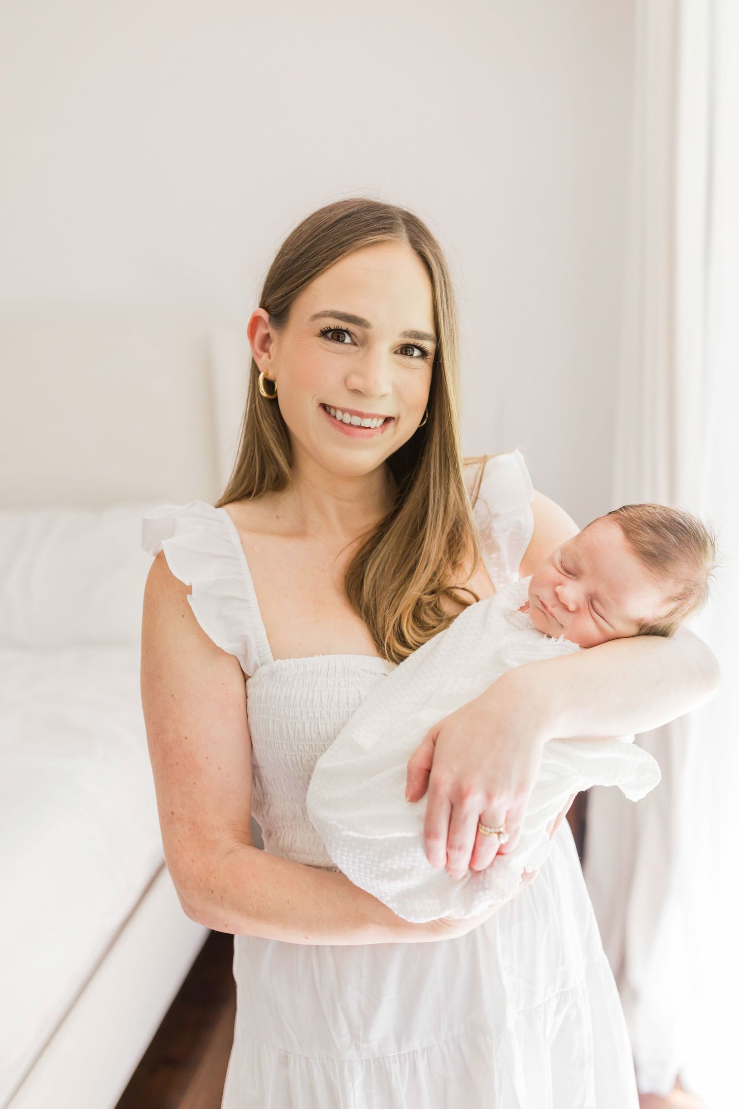 Woman in white dress holding sleeping newborn baby in bright room.