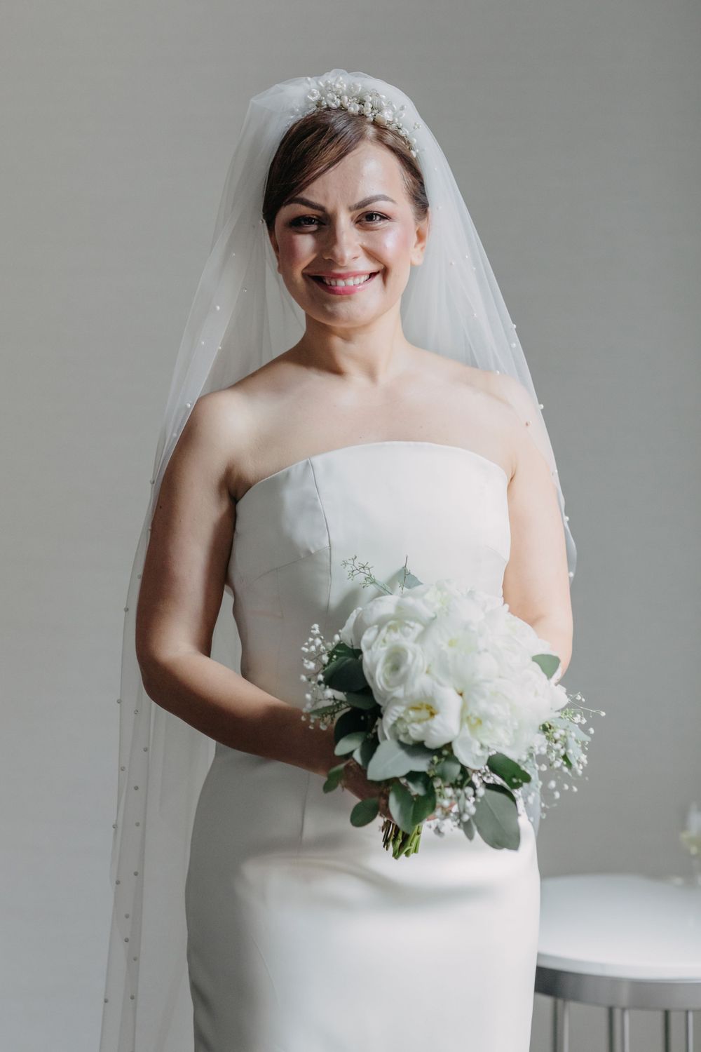 A bride in a strapless white wedding gown holds a bouquet of white flowers and greenery.