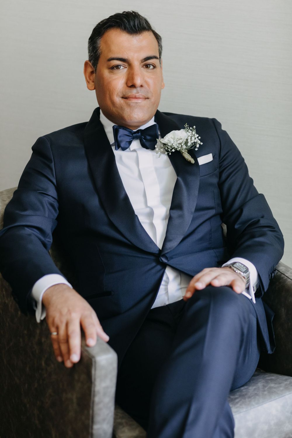 Groom in navy tuxedo and bow tie sits elegantly in chair while wearing boutonniere.