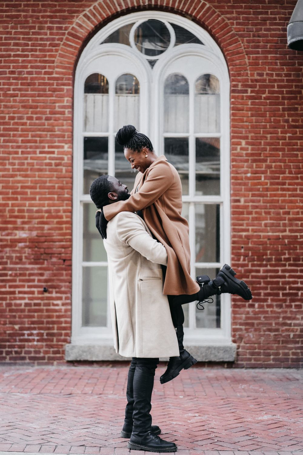 A loving couple embraces in front of an arched brick window, dressed in winter coats on a brick paved sidewalk, Georgetown , DC