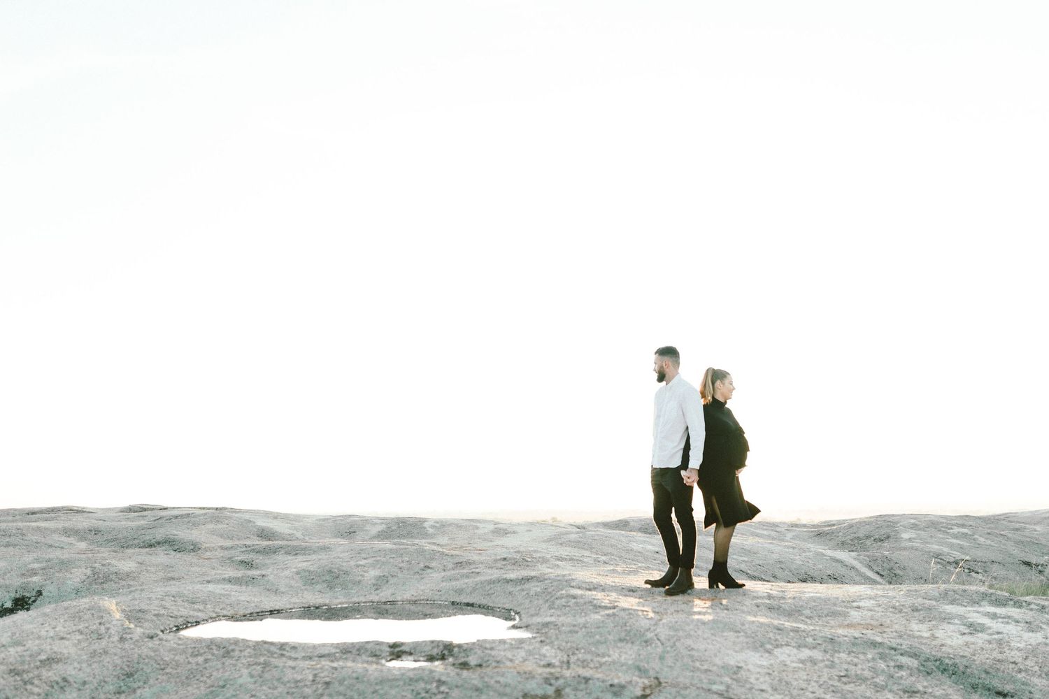 Couple stands on rocky outcrop with bright overexposed sky in background.