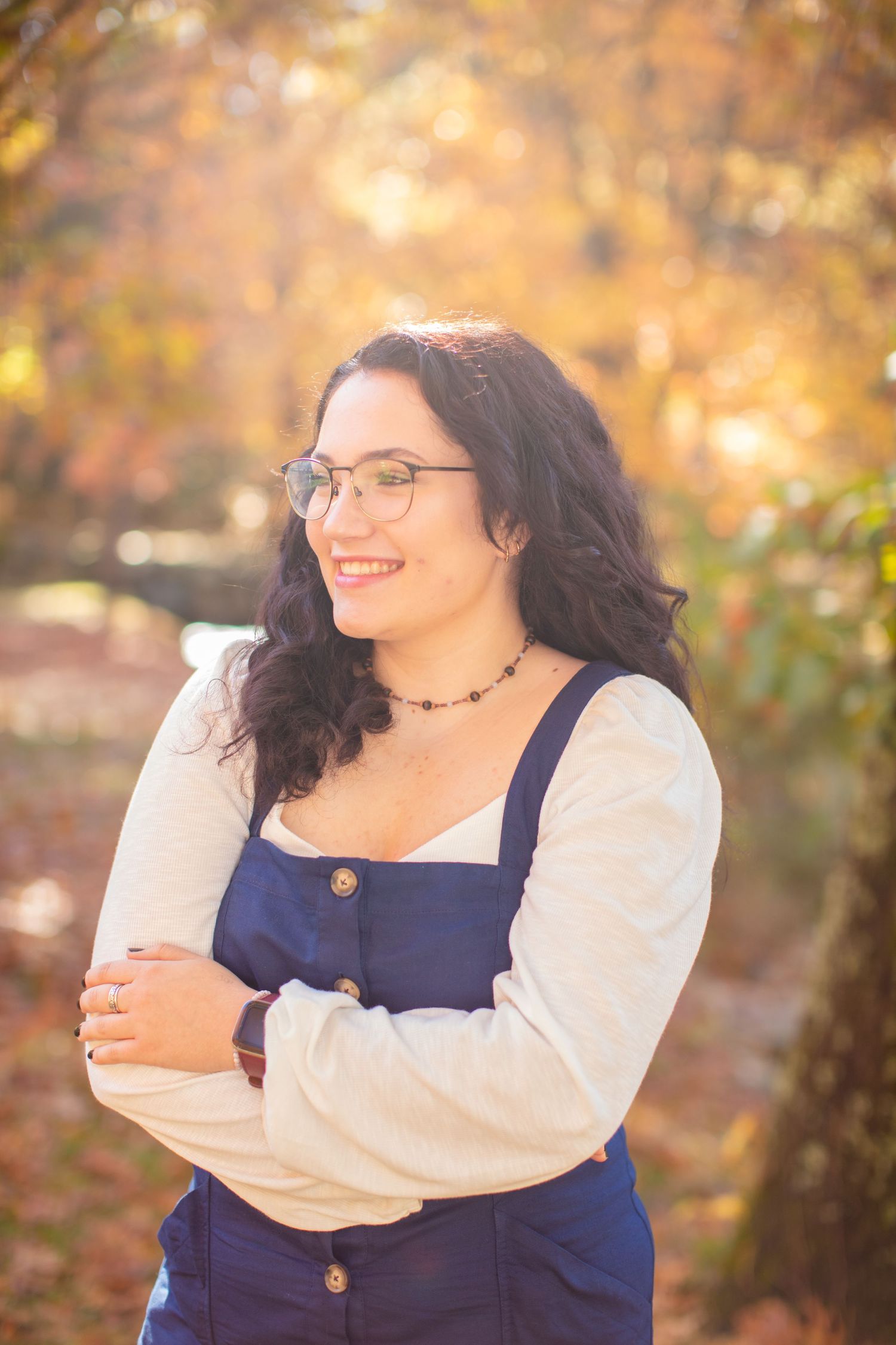 Portrait of person in blue overalls and white shirt smiling with autumn colors in background.
