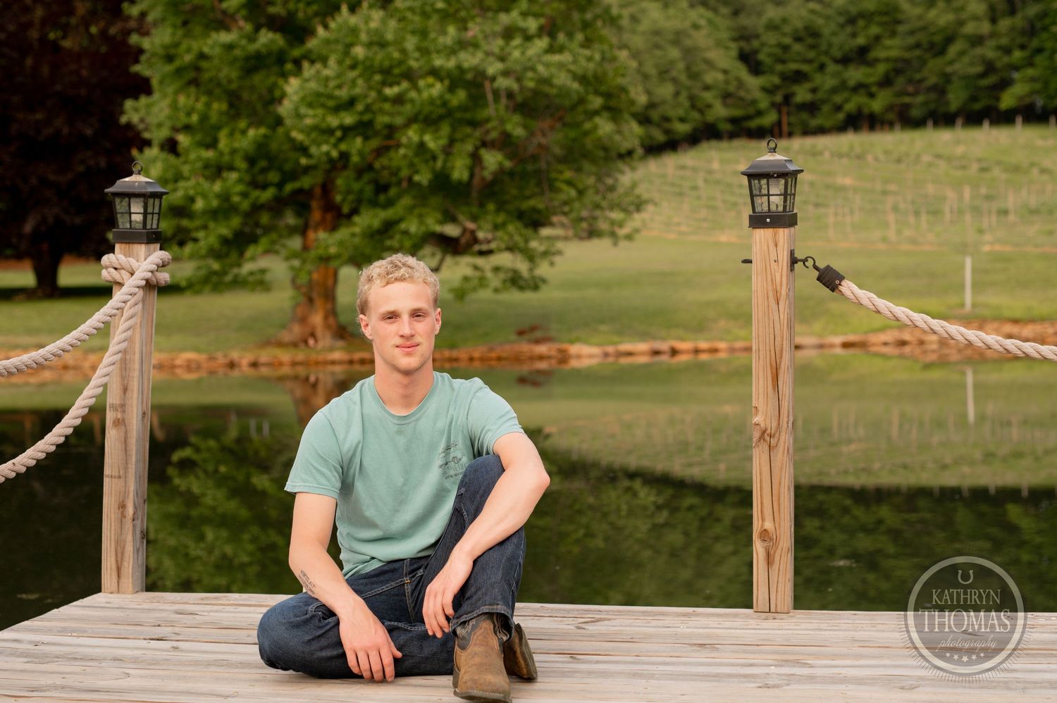 Person sits on wooden deck with rope railing overlooking peaceful pond.
