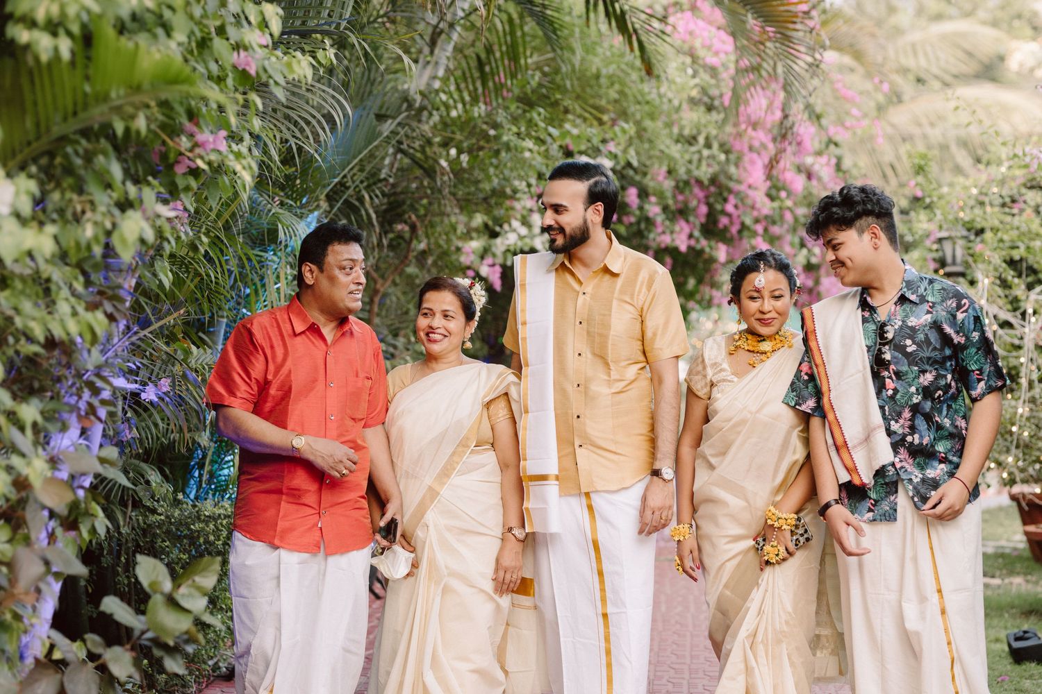 Wedding party poses together in coordinated cream and beige outfits under flowering trees.
