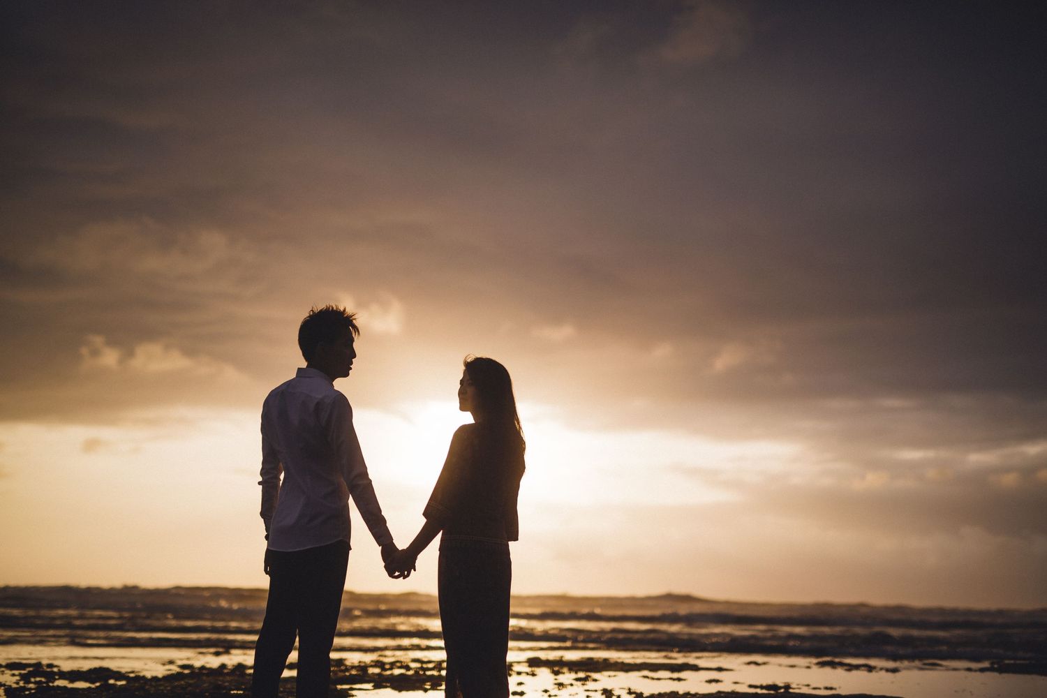Couple holding hands against moody sunset beach backdrop.