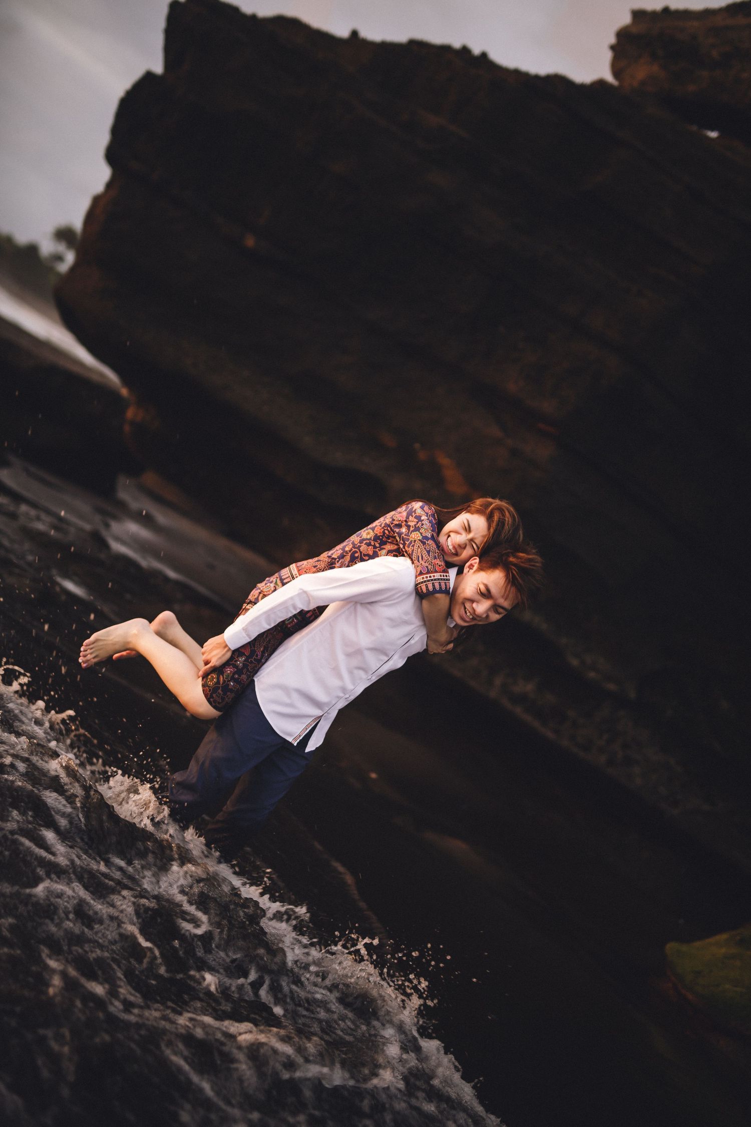 Playful couple splashing in ocean waves near rocky coastal cliffs at sunset.