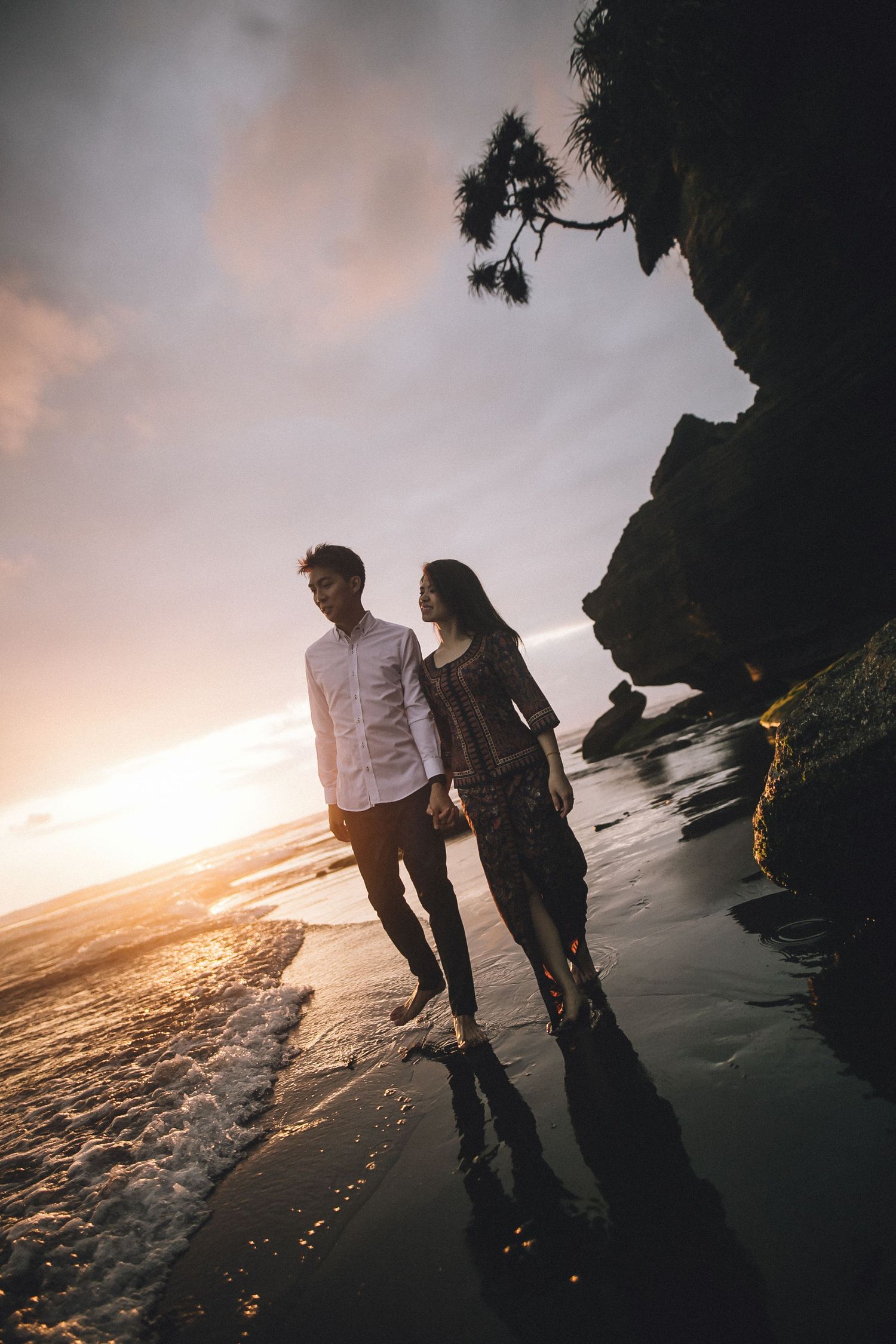Couple walking along dark sandy beach with dramatic cliffs and golden sunset.