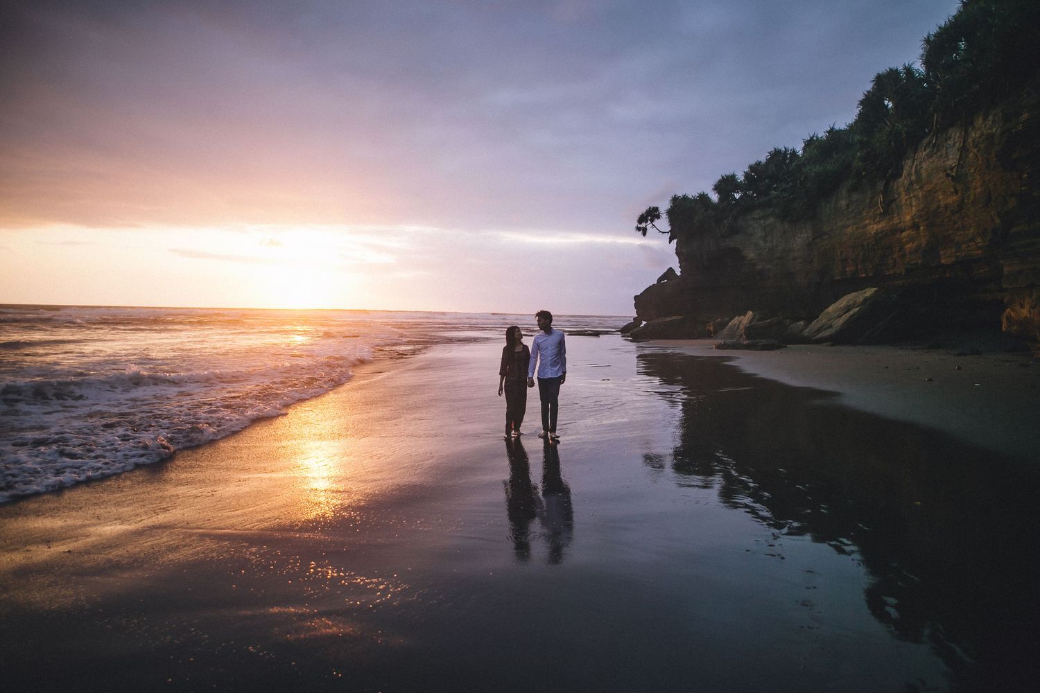 Silhouettes of a couple walking along a scenic beach at sunset with cliffs in the background.
