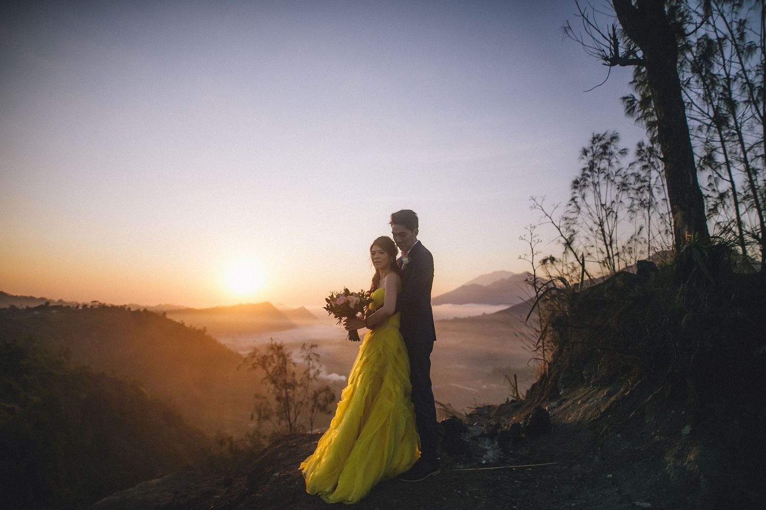 A couple in formal wear posing against a mountainous sunset backdrop.
